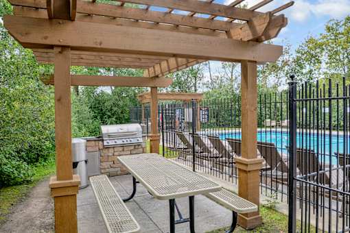 A wooden pergola with a grill and picnic table is situated next to a pool.