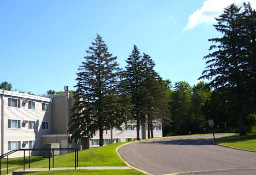 A road curves around a grassy area with trees and apartment buildings in the background.