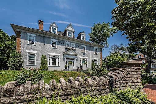 A large house with a grey facade and white trim is surrounded by a stone wall and greenery.
