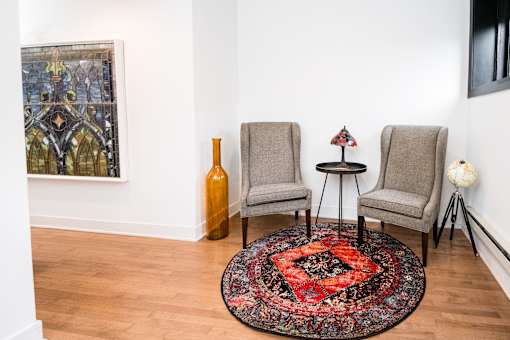A room with a large painting on the wall, two chairs, a table with a vase and a bottle on it, and a colorful rug on the floor.
