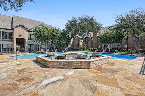 A pool surrounded by a stone patio with a fountain in the middle.