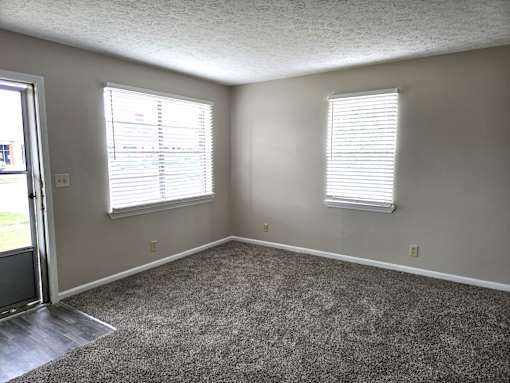 A room with a carpeted floor and two windows with blinds.