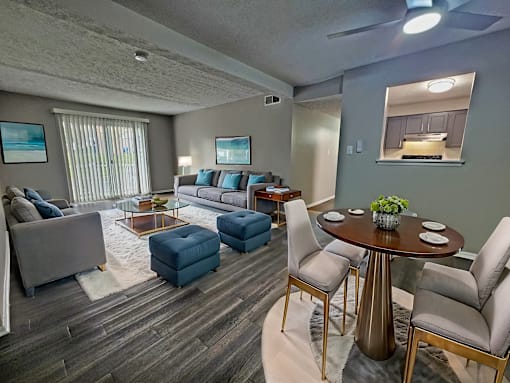 A living room with a grey couch, a blue ottoman, a glass coffee table, and a dining table with chairs.