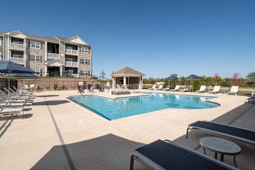 A large swimming pool surrounded by lounge chairs and umbrellas in front of a multi-story apartment building.