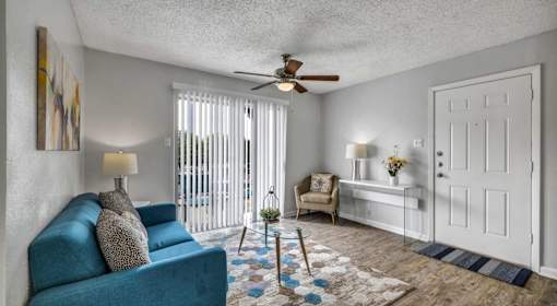 A blue couch sits in a living room with a ceiling fan and a window with blinds.
