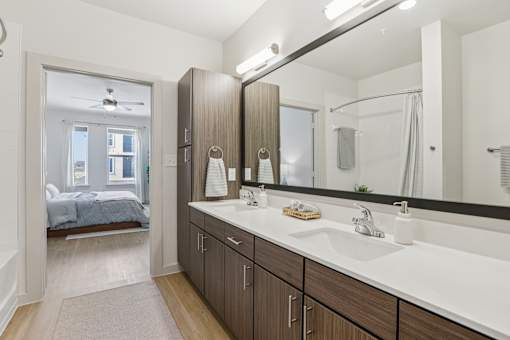 A bathroom with a large mirror and a bedroom with a bed and a window. at ThreeTwenty at the Vybe Apartments, Kyle, Texas