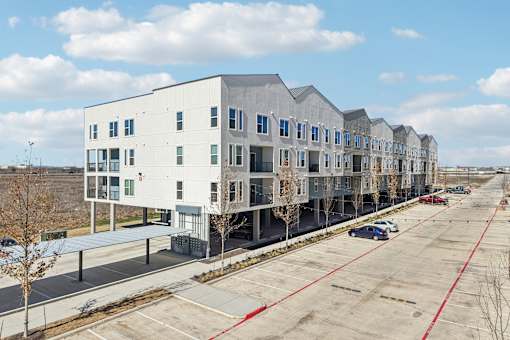 A parking lot in front of a building with a white and grey exterior. at ThreeTwenty at the Vybe Apartments, Kyle, 78640