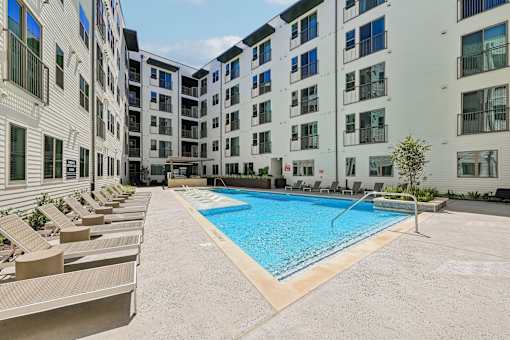 A swimming pool surrounded by sun loungers in front of apartment buildings.