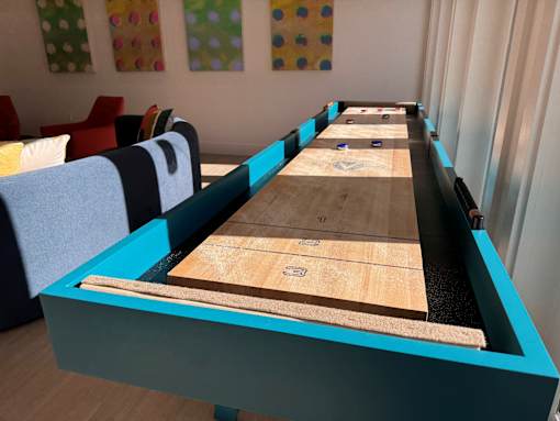 A shuffleboard table with blue and black trim is in the foreground of a room with colorful polka dot wall art. at ThreeTwenty at the Vybe Apartments, Texas, 78640