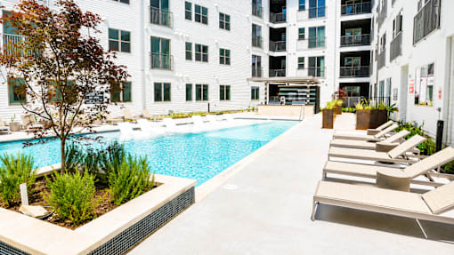 A pool surrounded by chairs and plants in front of a white building.