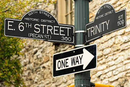 A street sign pole with signs for E 6th Street, Trinity Street, and a one way sign.