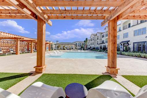 A wooden pergola over a pool with white chairs underneath.