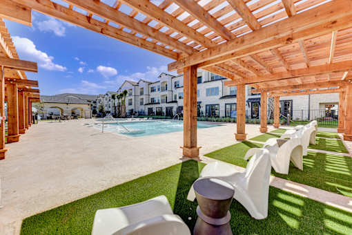 A wooden pergola over a patio with white chairs and a pool in the background.