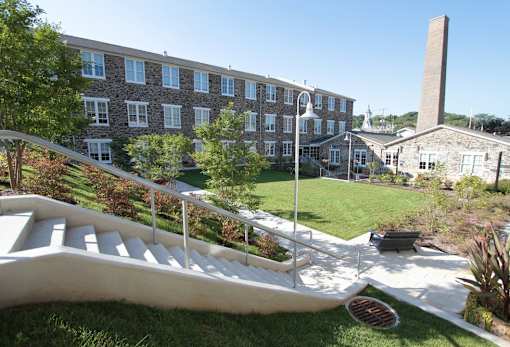 A building with a stone facade and a tall chimney is surrounded by a well-kept lawn and a white staircase.