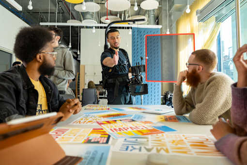 A group of people are sitting around a table with papers and pens, one person is standing and gesturing with their hands.