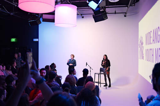 Two women are speaking to an audience in a dimly lit room.