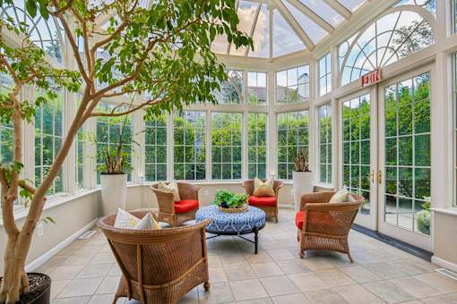 A sunroom with a glass roof and walls, furnished with a sofa, chairs, and a tree.