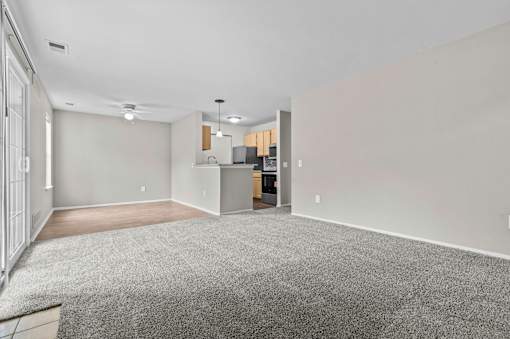 A spacious room with a grey carpet and a kitchen area in the background.