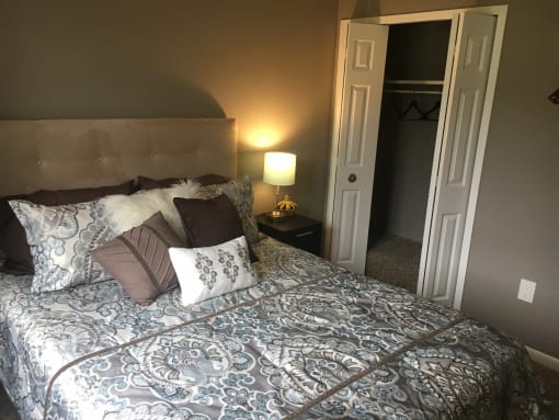 Bedroom with tan walls and a tan headboard. The bed is made in a blue patterned bed spread and white and brow accent pillows. Next to the bed is a lamp on a side table. The white closets doors are open to the right of the bed.