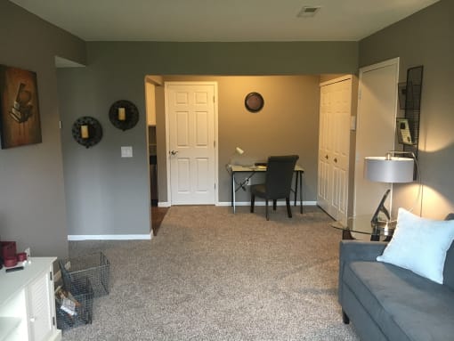 Living room with tan carpet and grey walls. A couch is up against the right wall. In the distance is a table and chair.