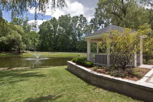 White gazebo to the right of photo with trees, greenery and lined with wood surround.  Green grass in the middle leading up to a pond with a fountain.  Sky and green trees in the background.