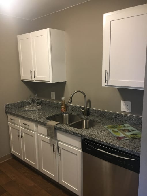 A kitchen with white cabinets and grey counter tops, grey walls, hardwood like flooring, and a stainless steal sink and dishwasher.