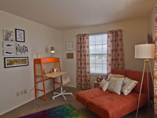 Spare bedroom with futon orange bed, lamp in right corner and red pattern curtains on window.