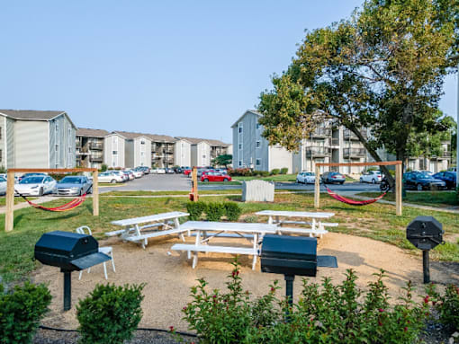 A park area with picnic tables and BBQ grills in front of apartment buildings.