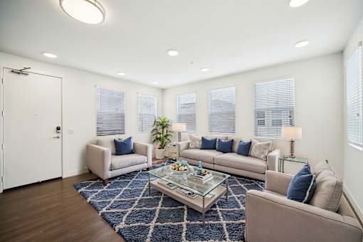 A living room with a white door, a grey couch, a blue rug, and a glass coffee table.