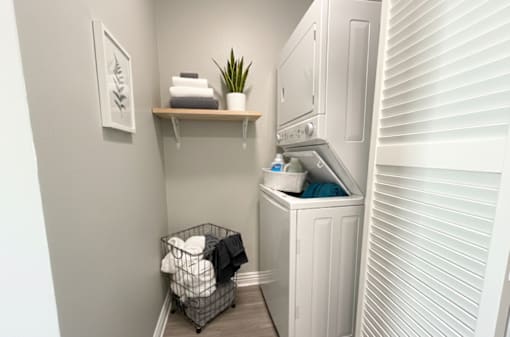 A small laundry room with a washer and dryer, a basket of clothes, and a shelf with a plant and towels.