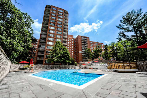 A swimming pool surrounded by a stone patio and trees with a red umbrella and chairs.