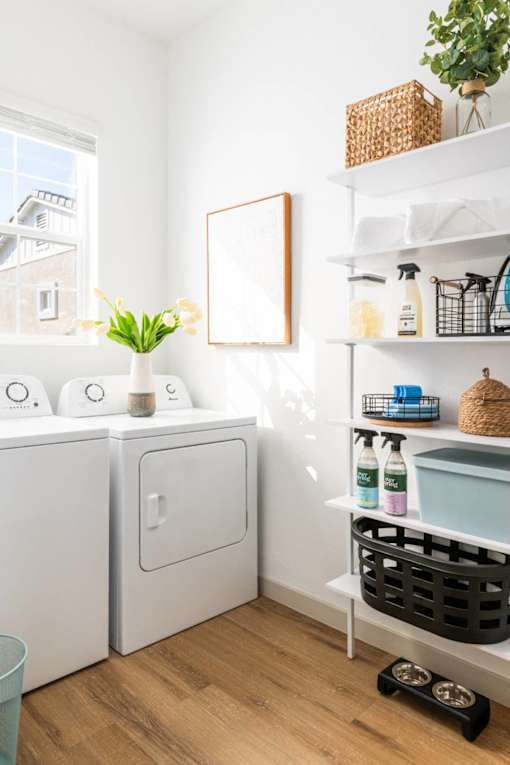 A white laundry room with a washer and dryer, a shelf with cleaning supplies, and a plant.