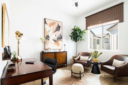 A living room with a brown chair, a black chair, a brown table, a white ottoman, a plant, and a painting on the wall.