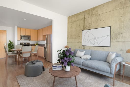 Apartments in Downtown San Jose, CA for Rent - Vendome Place - Open Floor Plan Living Area with Luxury Vinyl Tile Flooring, Area Rug, Exposed Concrete Accent Wall, Sofa, Ottoman, Round Coffee Table, and View of Kitchen and Dining Area.