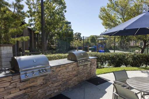 Apartments for Rent in Laguna Niguel CA - Pointe Niguel - BBQ Area with Two Grill Stations, Table, Chairs, Umbrella, Surrounding Trees, and View of Playground at Pointe Niguel, Laguna Niguel