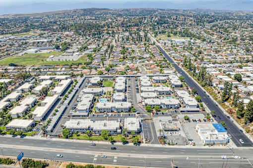 A large, open parking lot is surrounded by apartment buildings and a road with cars parked and driving.