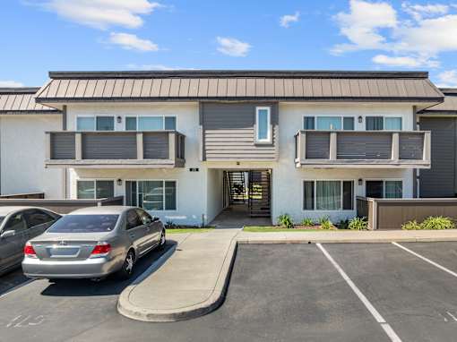 A silver car is parked in a parking lot in front of a two-story apartment building.