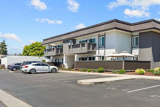 A parking lot in front of a grey and white building with a car parked in the lot.