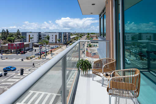A balcony with chairs and a potted plant overlooks a street with cars and buildings.