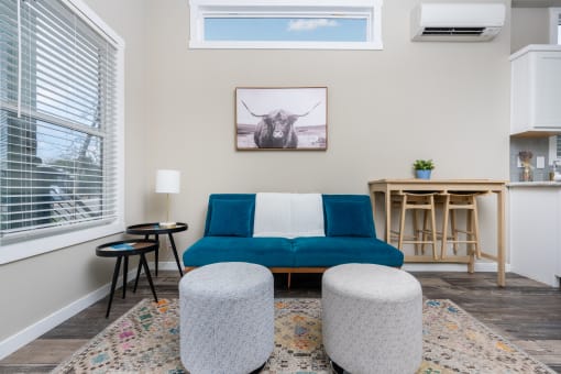 One-Bedroom Apartments in South Austin, TX - Casata Austin - Spacious Living Room with Wood-Style Flooring, Area Rug, Blue Couch, Round Ottomans, Side Tables with Lamp, Wooden Dining Table, AC Unit, Clerestory Window, and Large Window with Blinds.