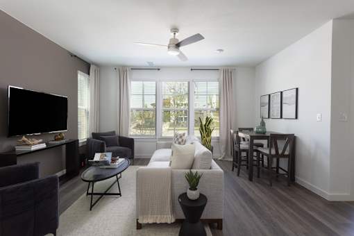 A living room with a grey couch, a black coffee table, a flat screen TV, a ceiling fan, and a dining table with chairs.