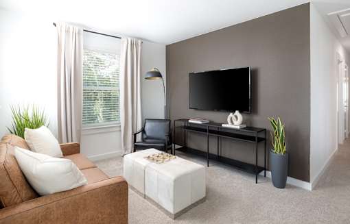 A living room with a brown couch, a white ottoman, a black chair, a flat screen TV, and a potted plant.