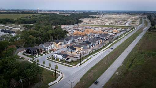 A road with a car driving on it and a residential area in the background.