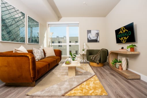 Apartments In South Park Los Angeles For Rent - Emerald - A Bright Living Space With A Fullsize Couch On One Wall And A Mounted TV On The Other Wall With Large Windows And Lightly Colored Walls