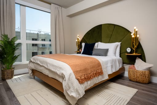 Pet-Friendly Apartments In South Park Los Angeles CA - Emerald - A Spacious And Bright Bedroom With A Full-Size Bed In The Center With One Wall Featuring A Nearly Floor-To-Ceiling Window, Wood-Style Flooring, Area Rug, And Lightly Colored Walls