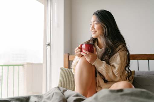 A woman is sitting on a bed holding a red mug.
