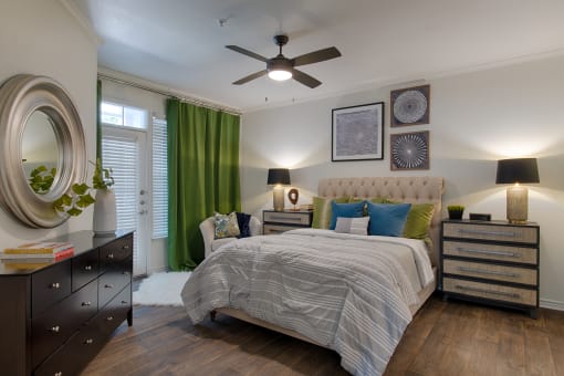 Apartments In Plano, TX - McDermott Place - Bedroom With Wood-style Flooring, A Large Window, An Exterior Glass Door With A Small Window Above, A Ceiling Fan, And Bedroom Furniture.