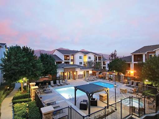 Pet-Friendly Apartments in Upland, CA - Park Central Apartments - Swimming Pool with a Spa, Sun Loungers, Lounge Seats, Pergola, and Umbrellas, Surrounded by a Metal Fence, Trees, and Landscaping.