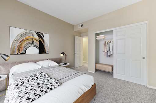 Dog-Friendly Apartments in Upland, CA - Park Central Apartments - Bedroom with Carpet Flooring, Large Bed, Nightstands, Lamps, Abstract Wall Art, and Oversized Closets with a Sliding Door.