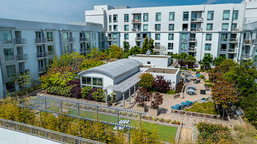 A modern building with a grey roof is surrounded by trees and other buildings.